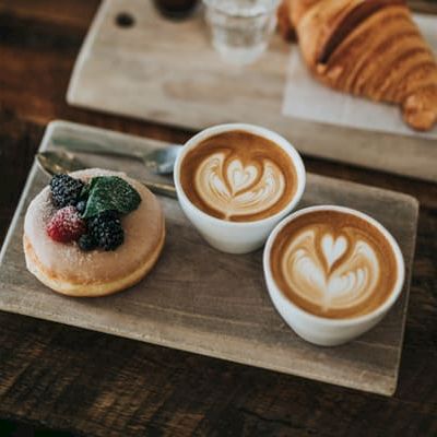 Two lattes with latte art hearts and a berry-topped pastry on a wooden tray, with a croissant in the background.