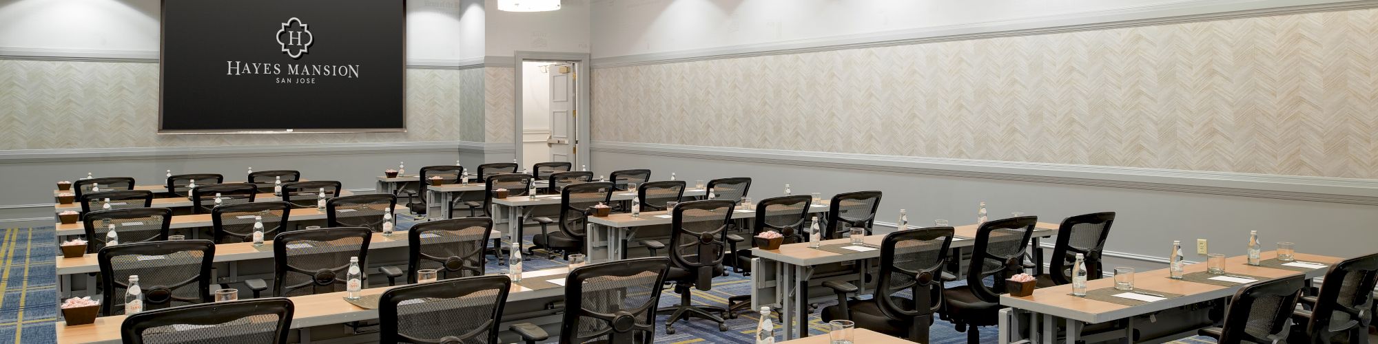 A modern conference room set up for a seminar or training, with rows of tables, black chairs, and a projector screen at the front.