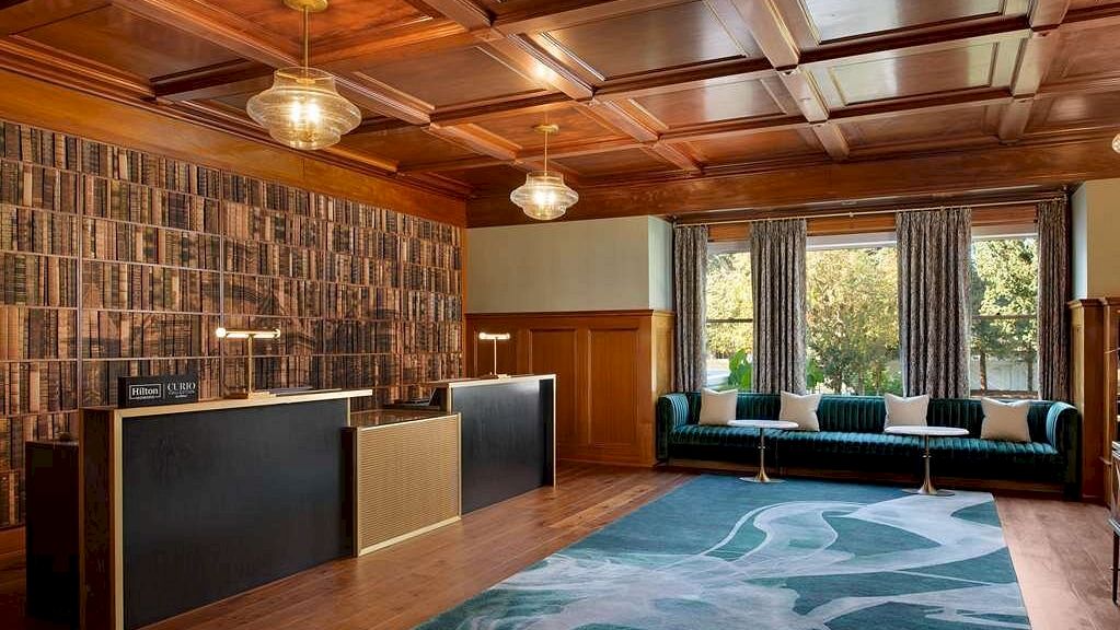 A stylish reception or lounge area with a large bookshelf wall, modern black-and-cream desks, warm wood ceiling, and a blue patterned rug by a windowed seating area.