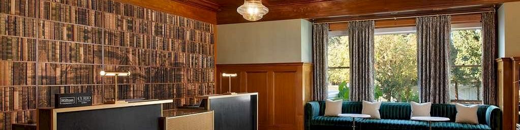 A stylish reception or lounge area with a large bookshelf wall, modern black-and-cream desks, warm wood ceiling, and a blue patterned rug by a windowed seating area.