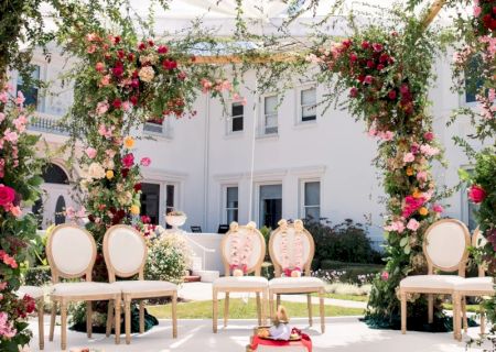 A garden wedding setup with a floral arch, elegant chairs, and a small red umbrella or decoration in the center, outdoor in a sunny courtyard.