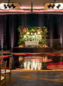 A wedding reception setup with a floral arch and stage, round tables with white linens and gold chairs, warm lighting, and a dance floor.