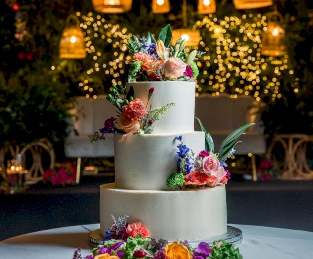 A stunning three-tier cake adorned with vibrant flowers, greenery, and soft lighting backdrop, perfect for a romantic celebration.