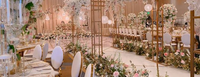 A lavish wedding reception setup with long tables, gold accents, white and pastel florals, elegant chairs, and lush floral arches creating an enchanting aisle.