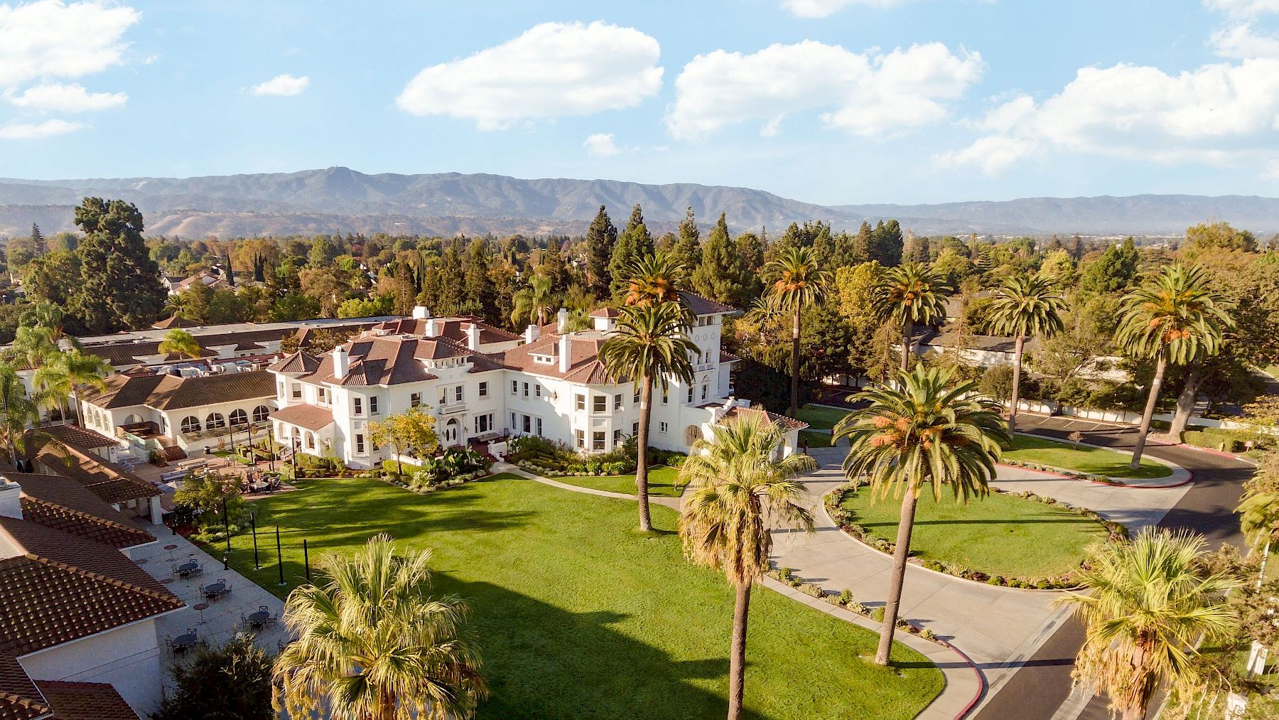 A sunny resort-style campus with white buildings, palm trees, green lawns, and winding paths under a blue sky with scattered clouds.