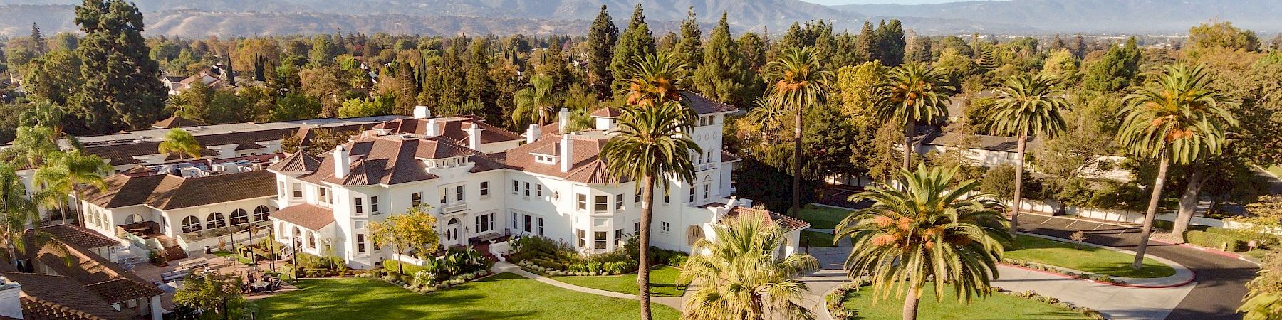 An aerial view of a sunny campus-like area with white buildings, palm trees, green lawns, and winding paths under a blue sky.