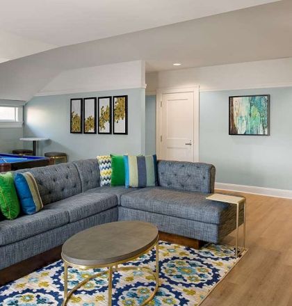 A bright, modern living room with a gray sectional, colorful cushions, a patterned rug, and a round coffee table, adjacent to a game area and pool table.