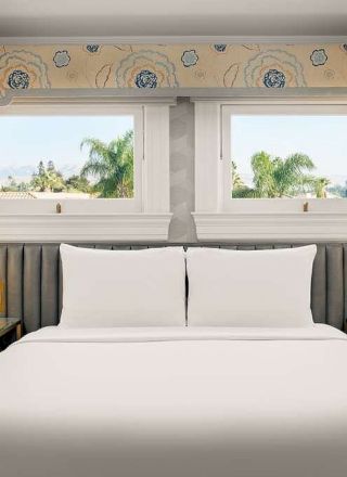 A neatly made hotel bedroom with a king bed, two nightstands with lamps, built-in shelves on either side, and a wide window view, ending with wallpapered walls and a valance.