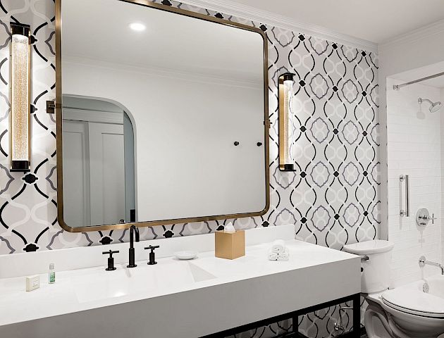 A modern bathroom with a large wall mirror, white double-sink vanity, patterned wallpaper, and a bright, airy feel.