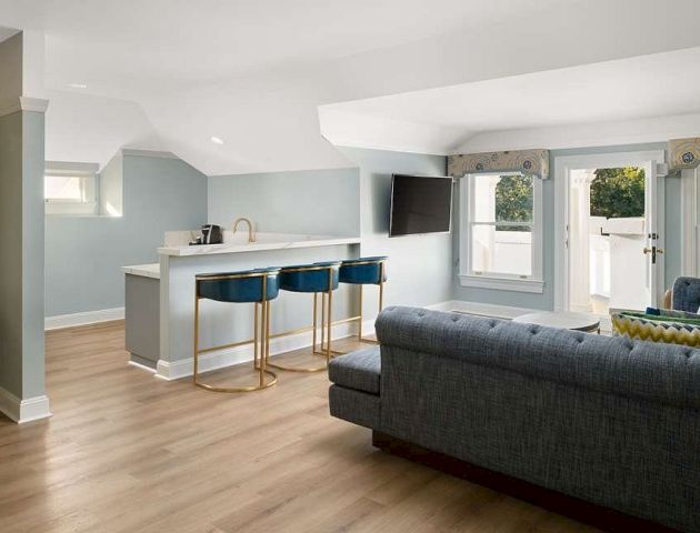 A bright, open living room with a gray sofa, white walls, large windows, hardwood floors, and a cozy, modern kitchen in the background.