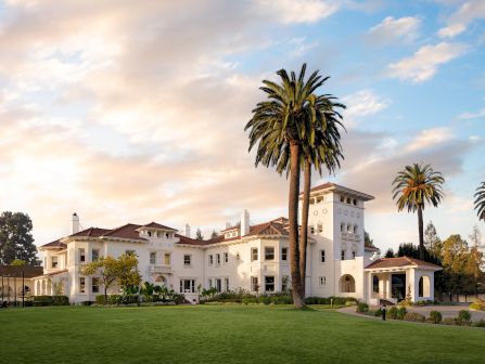 A white Mediterranean mansion with red-tile roof stands amid lush green lawns and tall palm trees under a pastel sunset sky.