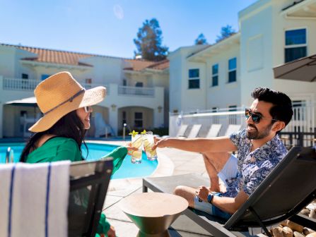 A couple lounges by a pool at a resort, toasting with drinks on a sunny patio.
