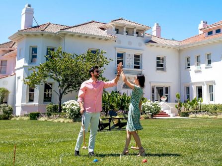 A man and woman high-five while playing on a green lawn in front of a white house with red-tile roofs and trees nearby.