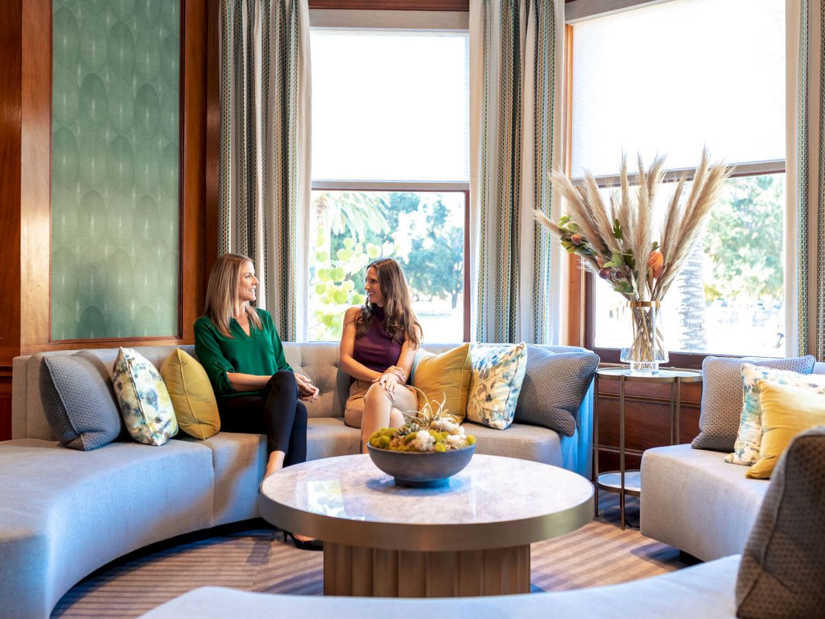 Two women sit and chat on a bright lounge sofa by large windows, with pillows, a coffee table, and a vase of flowers.